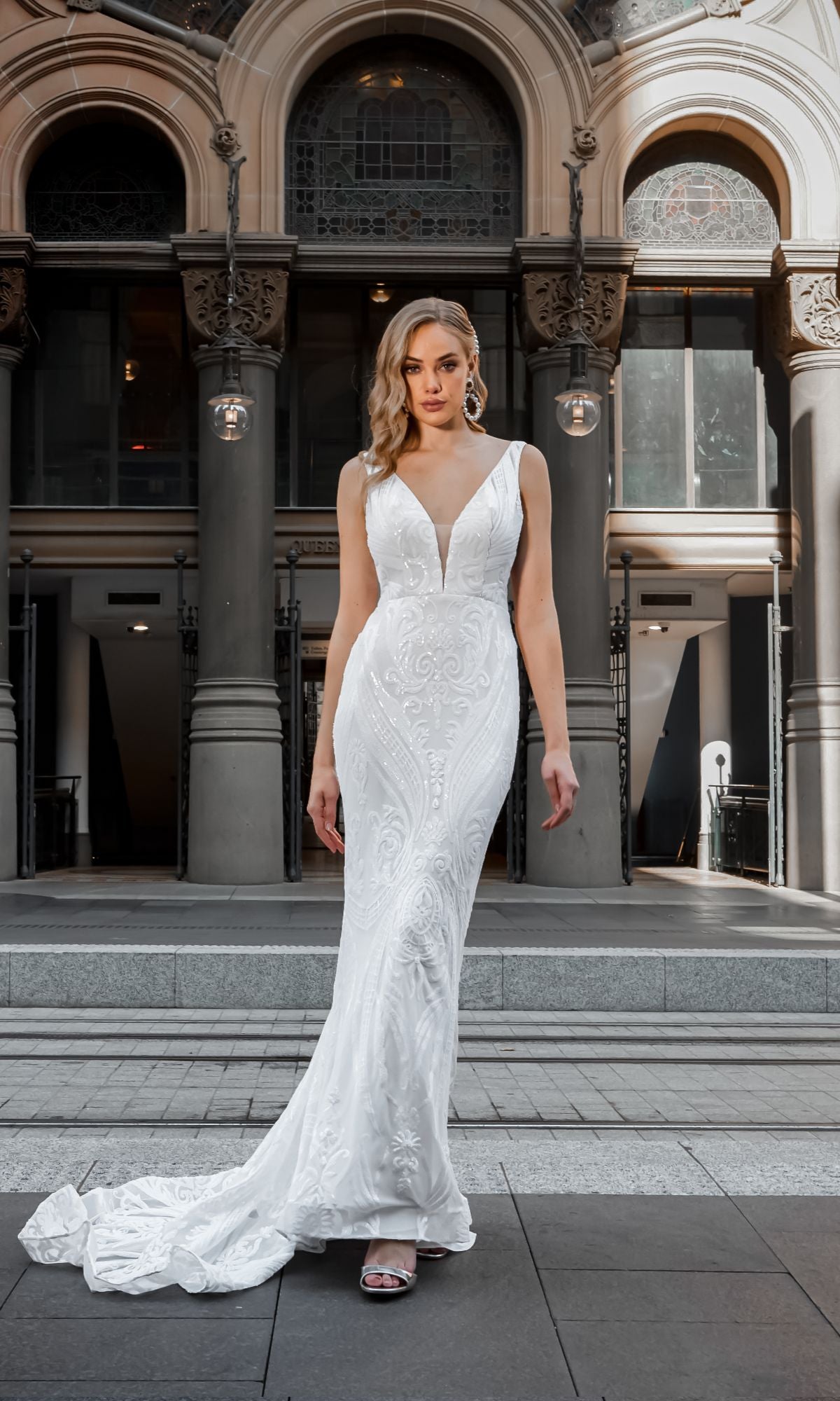 A woman wears the White Lace Mermaid Wedding Gown: Tina Holly BA109, featuring a long train, as she stands on stone steps before an ornate building.